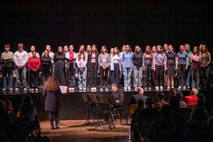 Ein großer Chor aus Jugendlichen steht auf einer Bühne und singt. Eine Frau dirigiert, während ein Pianist am Keyboard begleitet wird. Im Publikum sitzen weitere Personen.