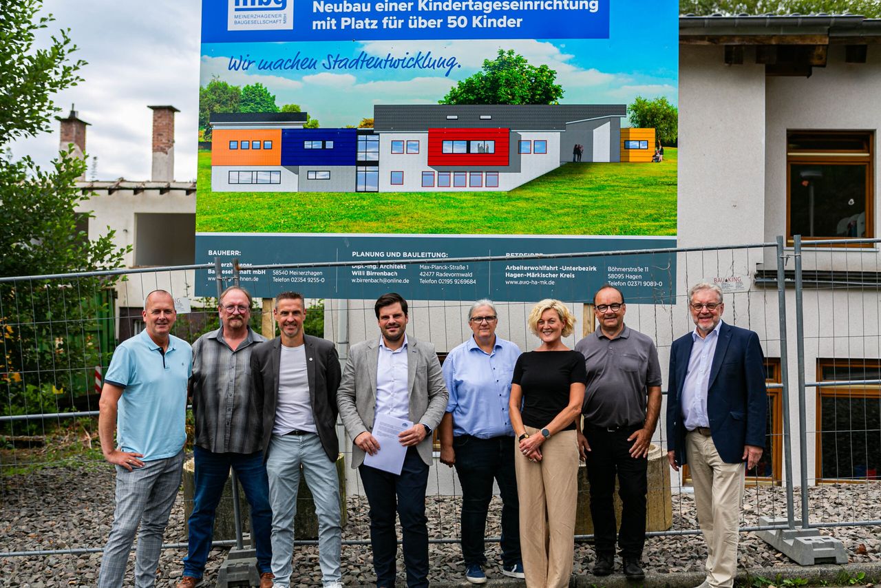 Eine Gruppe von neun Personen steht vor einer Baustellenplane, die eine Visualisierung eines neuen Kindergartens zeigt. Die Plane enthält Informationen zu Bauherr, Planung und Bauleitung sowie Kontaktinformationen. Die Personen sind größtenteils Männer in Anzügen und Hemden.