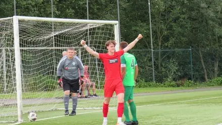 Ein Fußballspieler in roten Trikots jubelt mit erhobenen Armen nach einem Tor, während andere Spieler im Hintergrund stehen. Im Hintergrund ist ein Fußballtor und ein Zaun zu sehen.