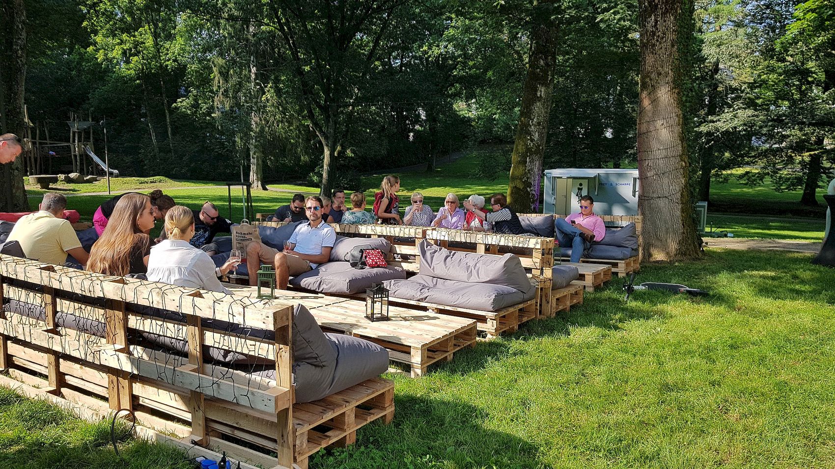 Menschen sitzen auf grauen Polstern auf Paletten-Sofas in einer Parkanlage unter Bäumen.
