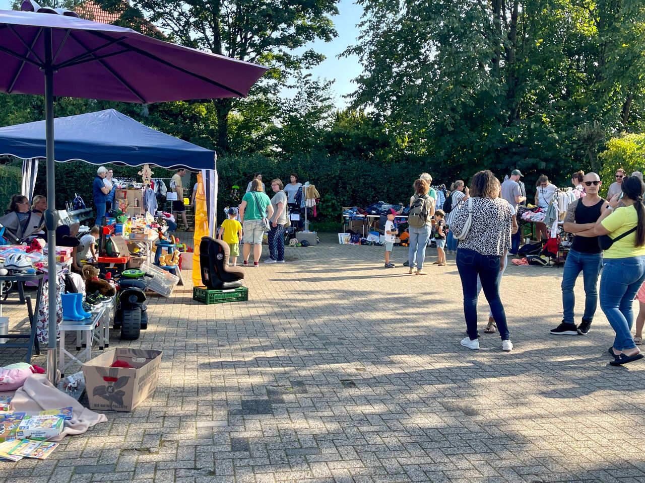 Menschen stöbern auf einem Gemeindefest an Verkaufsständen mit Spielzeug und Kleidung unter einem blauen Zelt und einem lila Sonnenschirm.