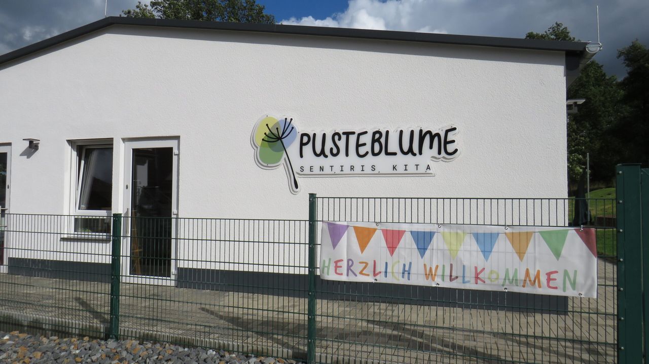 Weißer Gebäudeteil mit dem Kita-Logo „Pusteblume, Sentiris, KITA“ und einem Banner mit der Aufschrift „Herzlich Willkommen“.