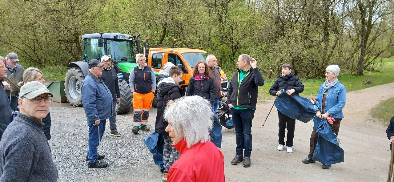 Eine Gruppe von etwa 15 Personen steht auf einem Kiesweg vor einem blauen Traktor und einem orangefarbenen Fahrzeug. Sie scheinen sich zu unterhalten, einige tragen Warnwesten und Müllsäcke.