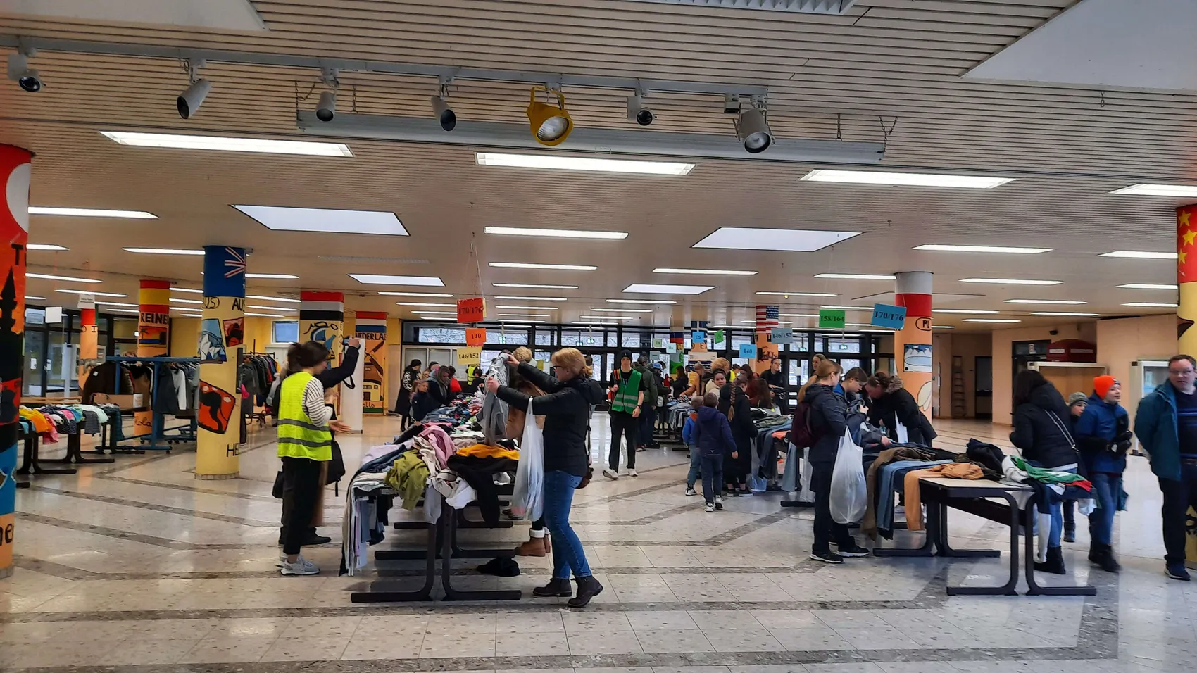 Besucher durchstöbern Tische mit Kleidung in einer großen Halle während eines Kinderkleiderbasars.