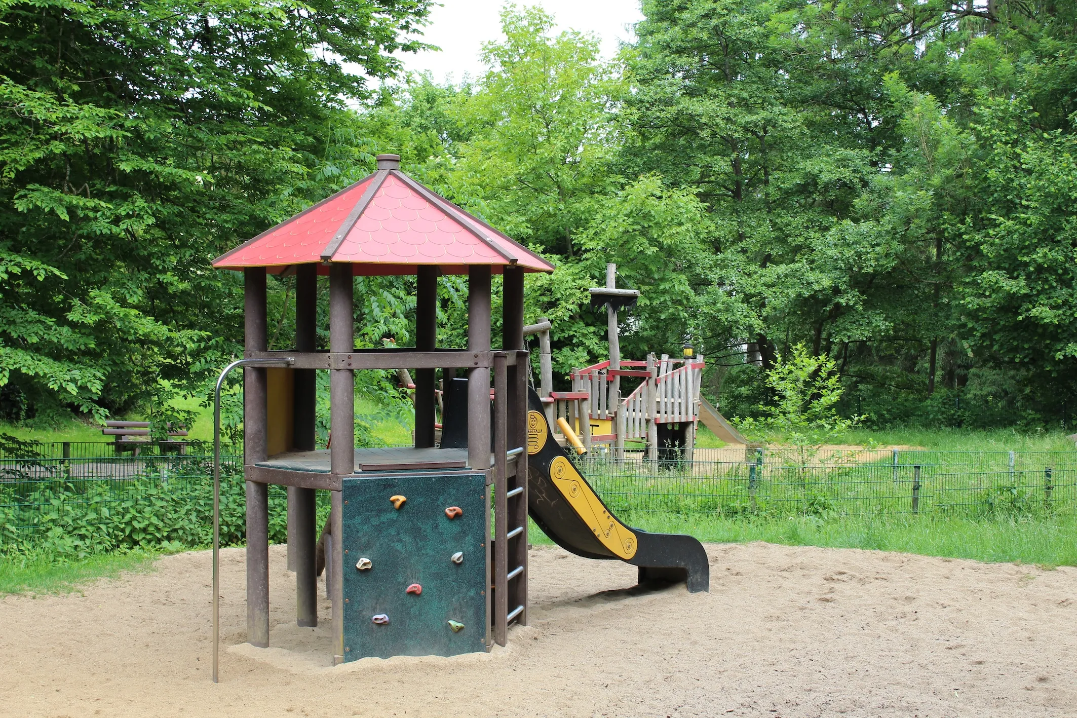 Spielturm mit Kletterwand und Rutsche auf einem Sandplatz im Hülsenbecker Tal, umgeben von Bäumen und einem Zaun.