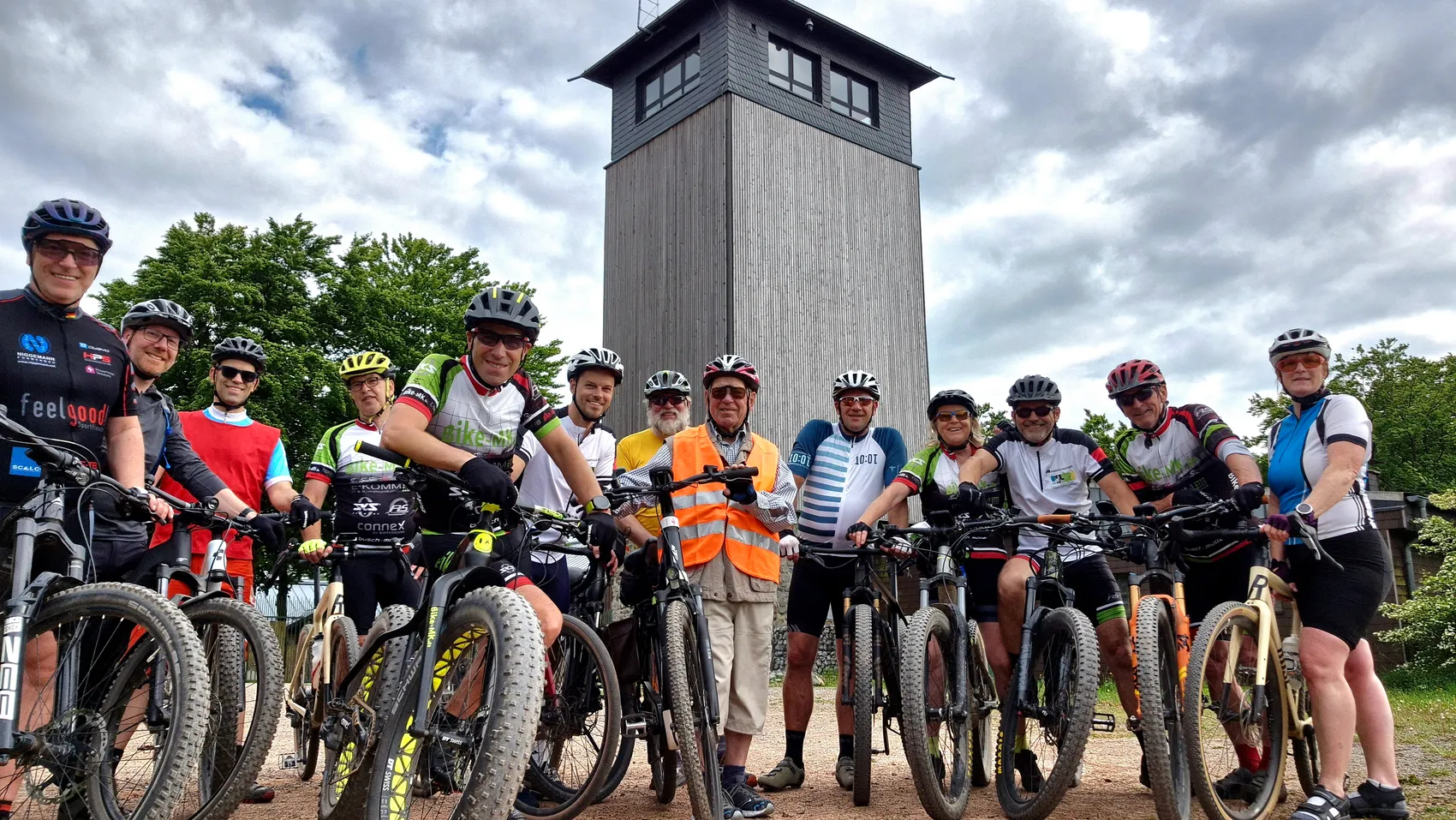 Eine Gruppe von Radfahrern mit Helmen und Fahrrädern steht vor einem hohen, grauen Aussichtsturm in Herscheid, um die Teilnahme an der Stadtradeln-Aktion zu zeigen.