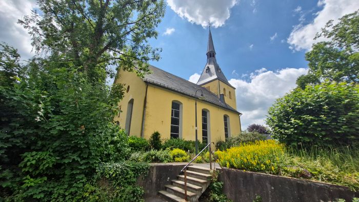 Gelbe Kirche mit schlankem Turm und steilem Dach in Rönsahl, umgeben von Bäumen und Büschen. Eine Steintreppe führt zum Eingang.