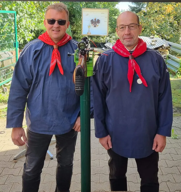 Martin Abel und Mike Braumann in blauen Hemden und roten Halstüchern stehen neben einem auf einem grünen Ständer montierten Gewehr.
