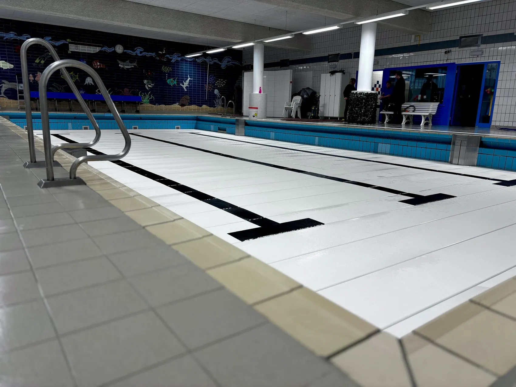 Leeres Schwimmbecken mit blauen Bahnen und weiß gefliestem Beckenrand. Im Hintergrund eine Wand mit Unterwasserillustrationen und ein Bereich mit Sitzgelegenheiten und Personen.