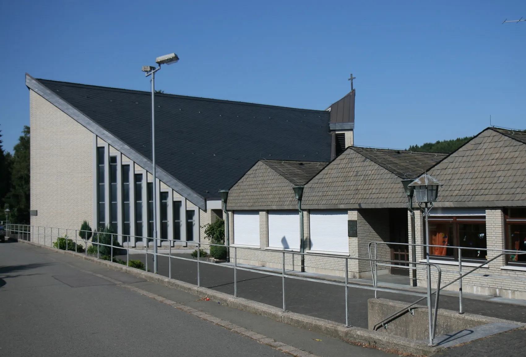 Moderner Kirchenbau mit Satteldach und schlanken Fensterreihen, daneben ein niedrigerer Anbau mit Walmdach und Kreuz.