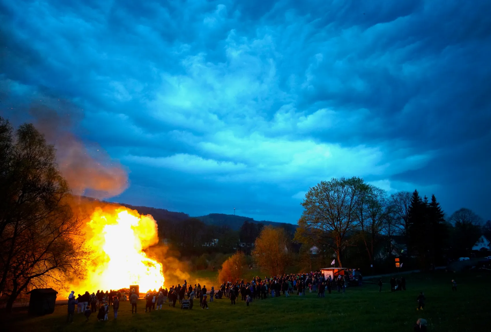 Großes Osterfeuer mit vielen Menschen im Kreis auf einer Wiese bei Dunkelheit, im Hintergrund Hügel und ein bewölkter Himmel.