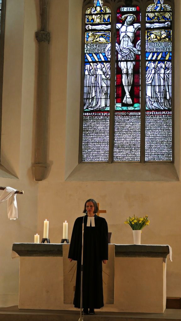 Pfarrerin Christine Rosner steht vor dem Altar in der Christuskirche Plettenberg und hält eine Ansprache. Im Hintergrund ein großes Buntglasfenster mit religiösen Motiven.