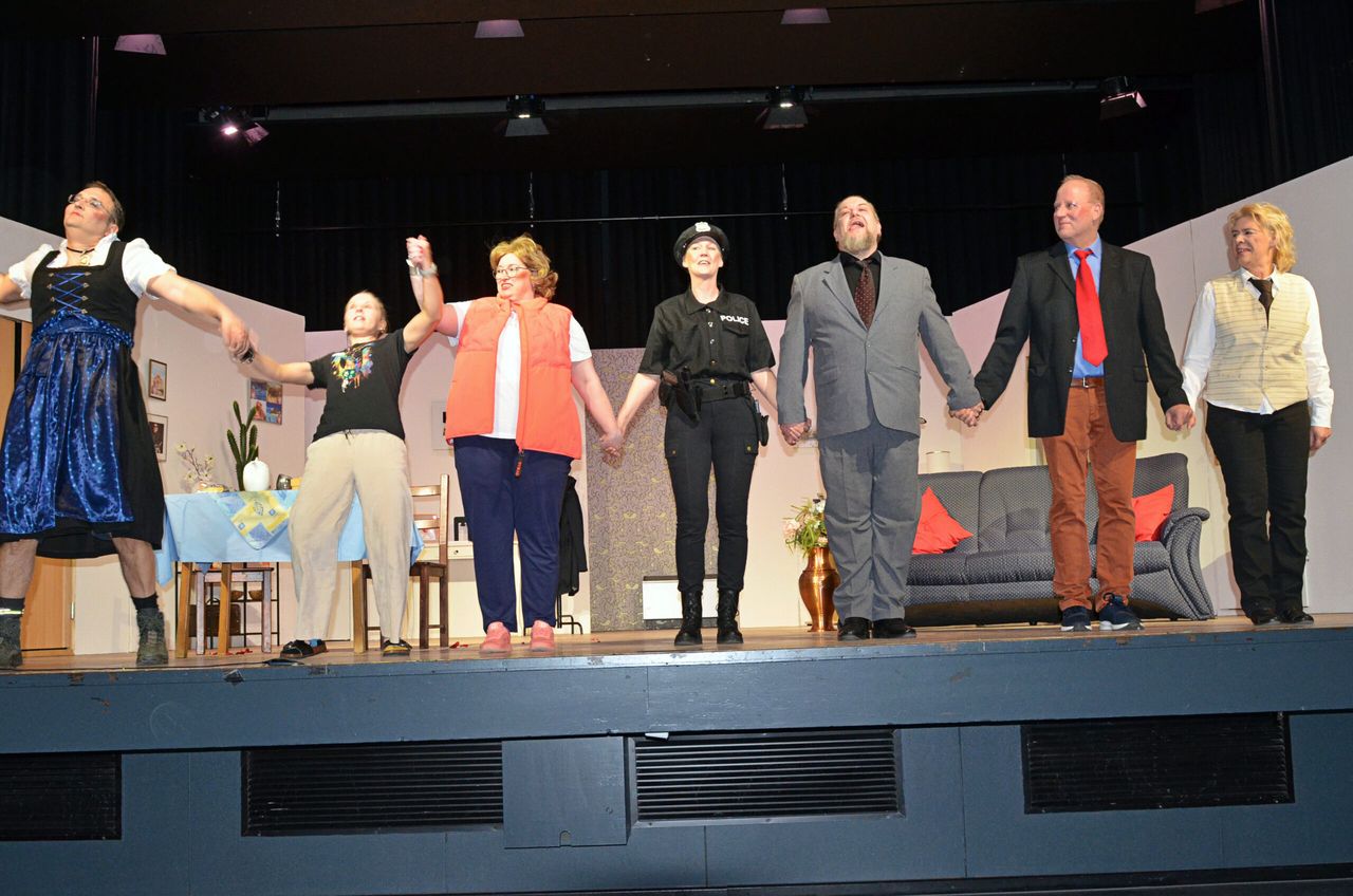 Sieben Darstellerinnen und Darsteller halten sich auf einer Bühne an den Händen und blicken ins Publikum. Im Hintergrund steht ein Sofa und eine gedeckte Tafel.