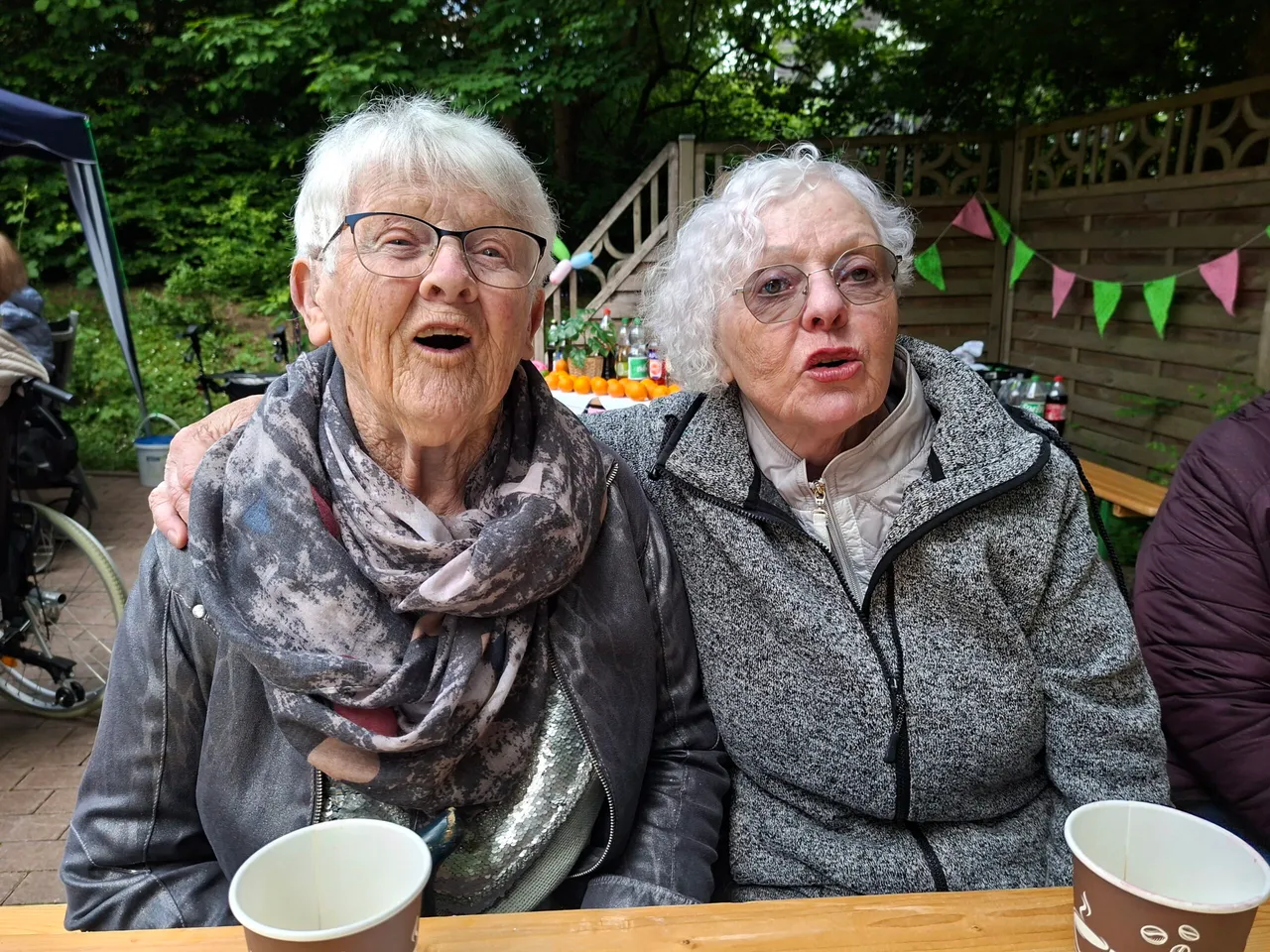 Zwei ältere Frauen sitzen eng nebeneinander an einem Tisch, beide schauen überrascht in die Kamera. Im Hintergrund ein festlich geschmückter Garten.