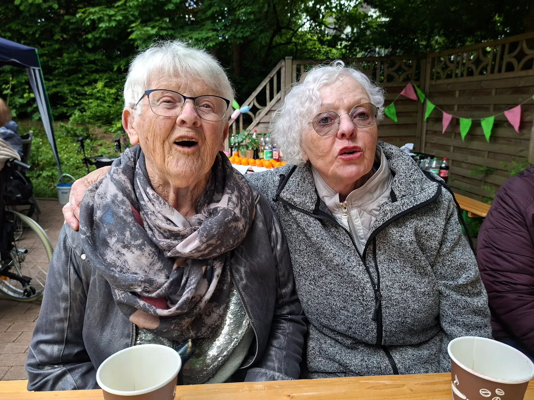 Zwei ältere Frauen sitzen eng nebeneinander an einem Tisch, beide schauen überrascht in die Kamera. Im Hintergrund ein festlich geschmückter Garten.