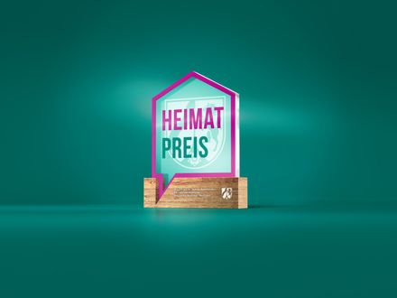 Ein gläserner Pokal in Form eines Hauses mit dem Schriftzug „HEIMATPREIS“ in Pink und Weiß. Darunter ein Holzsockel mit dem Text „Ministerium für Heimat, Kommunales, Bau und Digitalisierung des Landes Nordrhein-Westfalen“ und einem Logo.