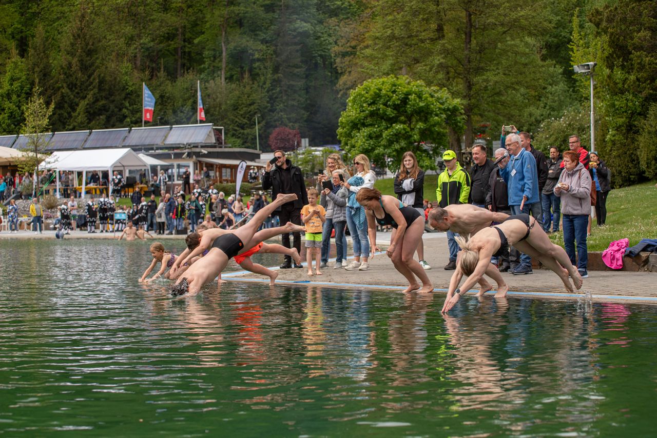 Mehrere Schwimmer springen gleichzeitig von einer Betonmauer in ein Becken, während Zuschauer am Ufer stehen.