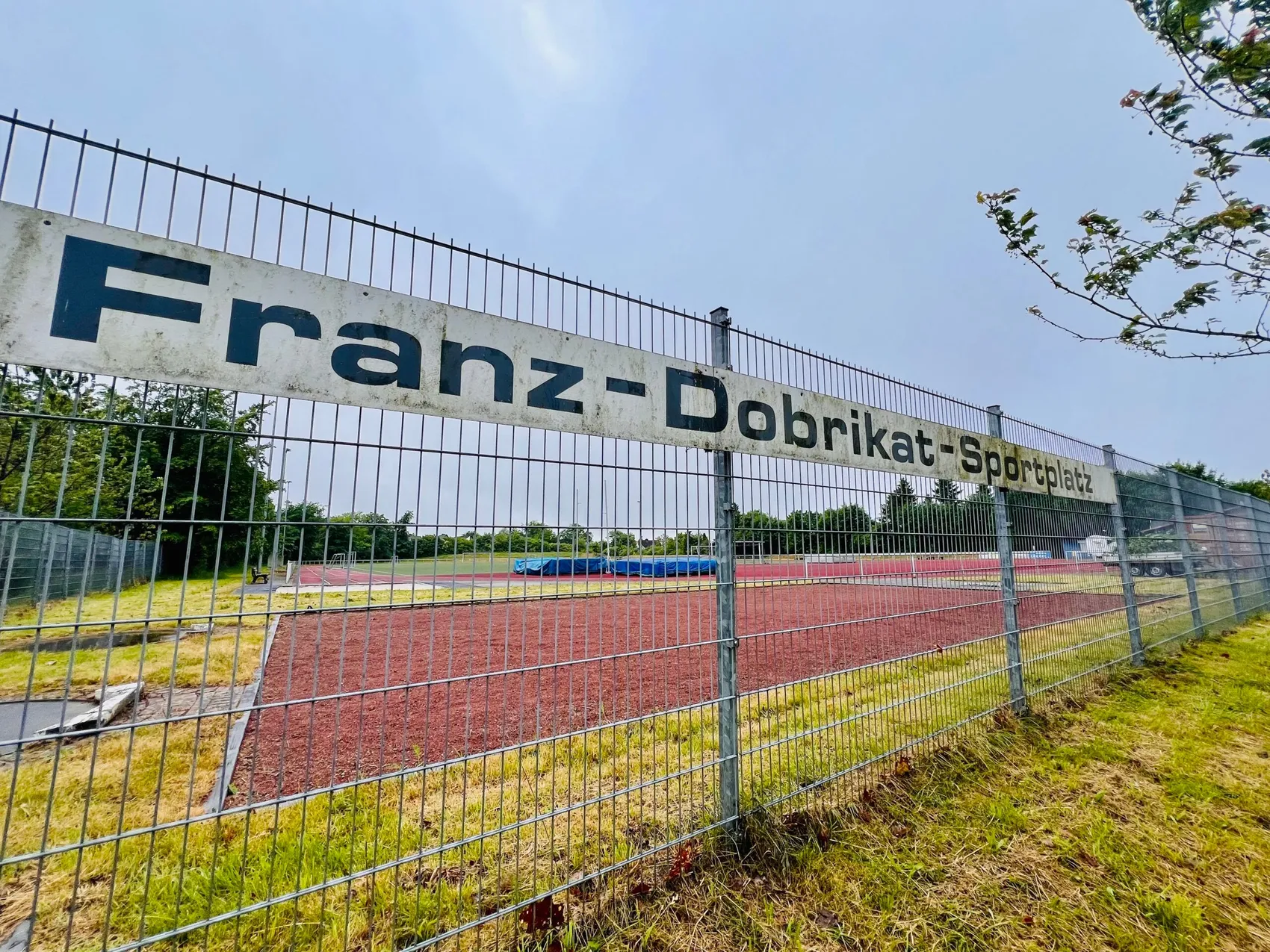 Schild „Franz-Dobrikat-Sportplatz“ über einem roten Tartanbahnlaufplatz mit grüner Rasenfläche im Hintergrund.