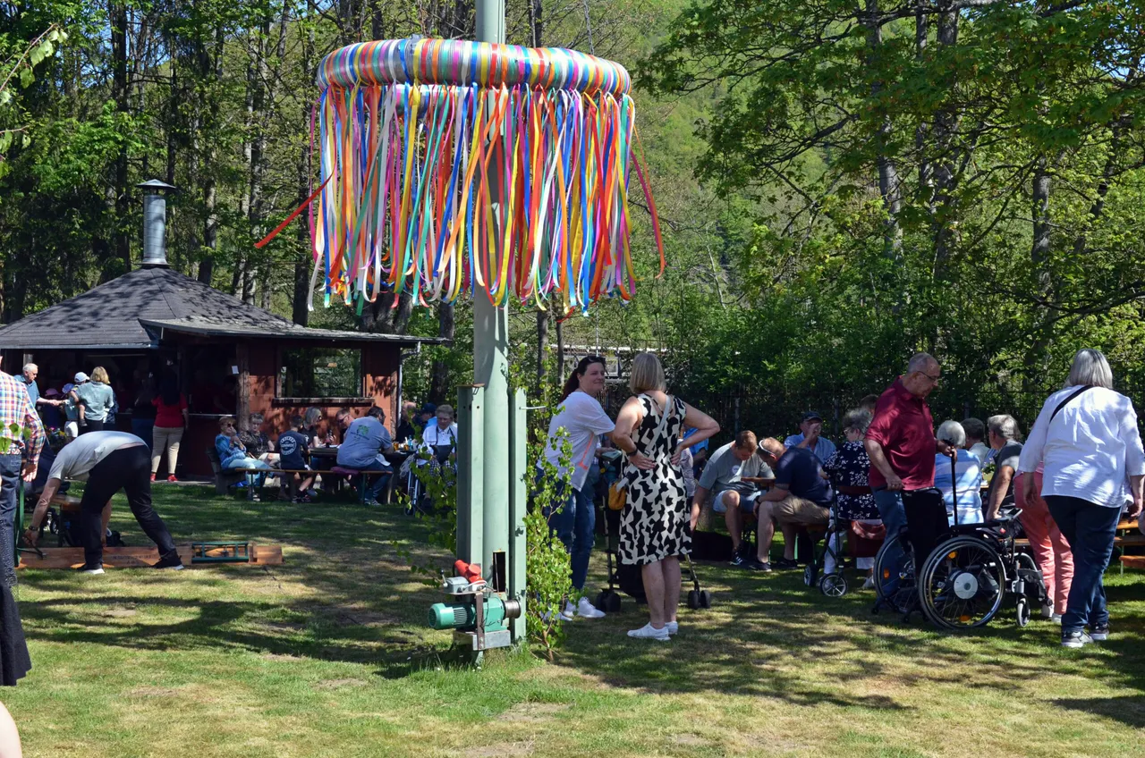 Menschen feiern im Freien, einige sitzen an Tischen, andere stehen und unterhalten sich. Im Vordergrund ein mit bunten Bändern geschmückter Mast.