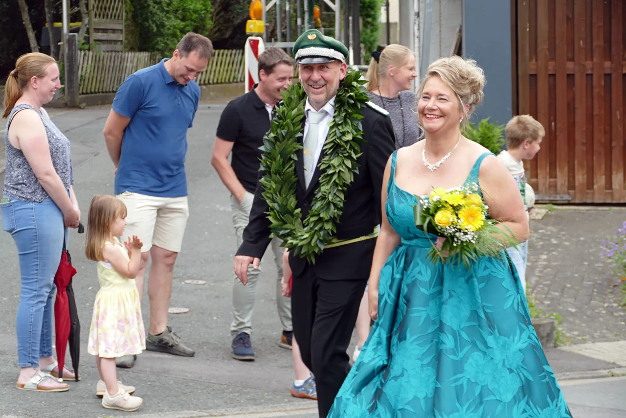 Ein Mann in Uniform und eine Frau in einem türkisfarbenen Kleid lächeln und gehen im Rahmen eines Umzugs nebeneinander. Um den Hals des Mannes hängt ein Kranz, die Frau trägt einen Blumenstrauß.