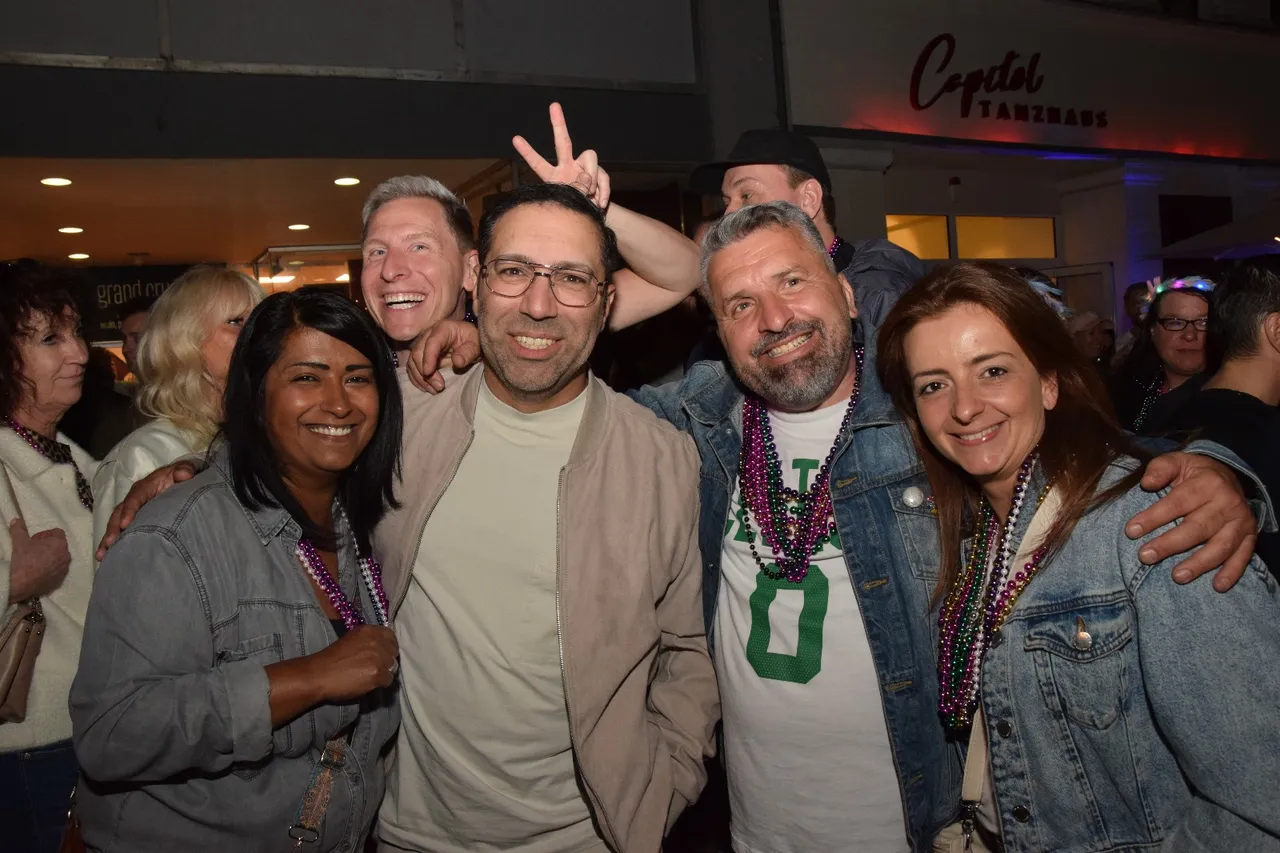 Eine Gruppe von Menschen lächelt in die Kamera, einige tragen Perlenketten und machen das Victory-Zeichen. Im Hintergrund ist ein Gebäude mit der Aufschrift „Capitol Tanzhaus“ zu sehen.