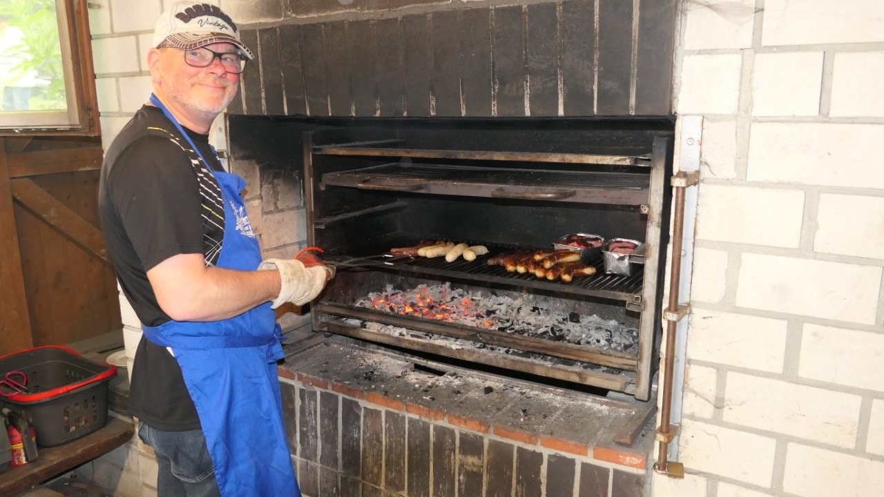 Ein Mann mit Schürze und Handschuhen grillt Würstchen und Fleischspieße in einem großen gemauerten Grill.
