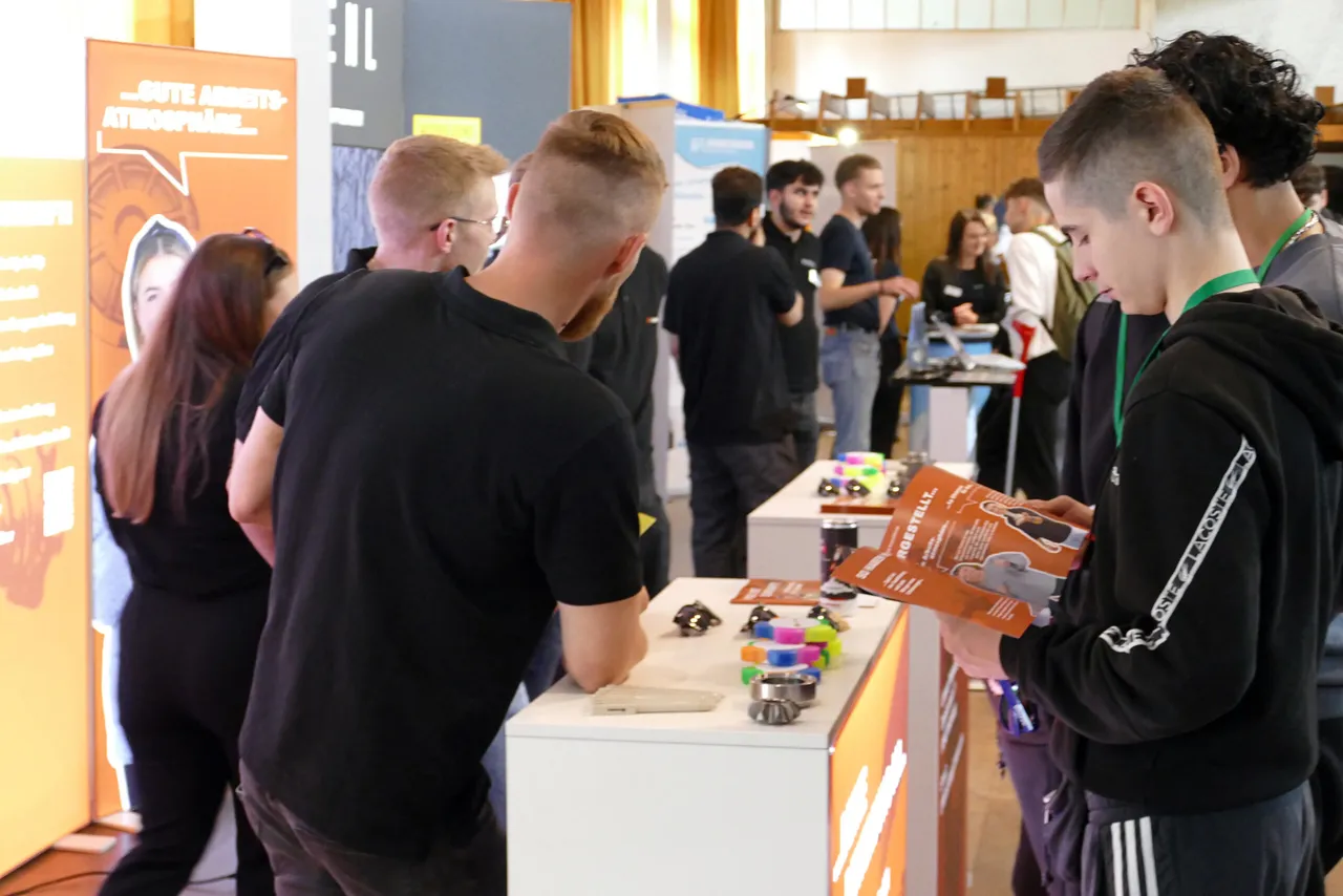 Zwei junge Männer betrachten an einem Messestand verschiedene Ausbildungsmodelle und eine Broschüre. Im Hintergrund sind weitere Besucher und Stände zu sehen.