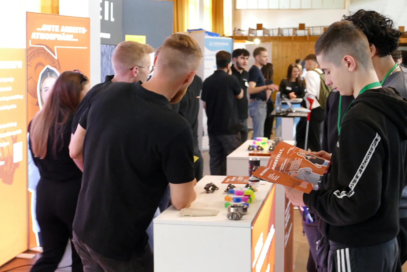 Zwei junge Männer betrachten an einem Messestand verschiedene Ausbildungsmodelle und eine Broschüre. Im Hintergrund sind weitere Besucher und Stände zu sehen.