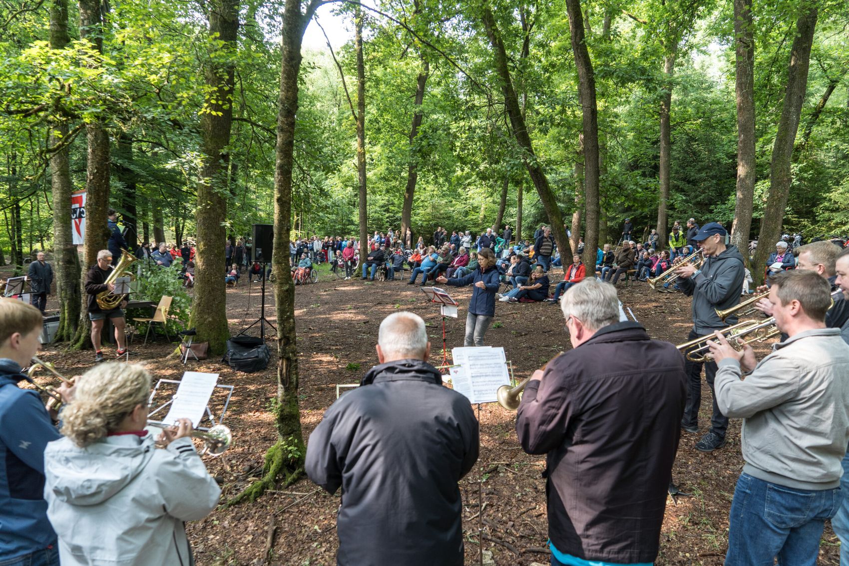 Bläser spielen im Wald vor einer großen Menschenmenge, die auf Stühlen und Bänken sitzt. Im Hintergrund stehen Bäume und ein rotes Banner.