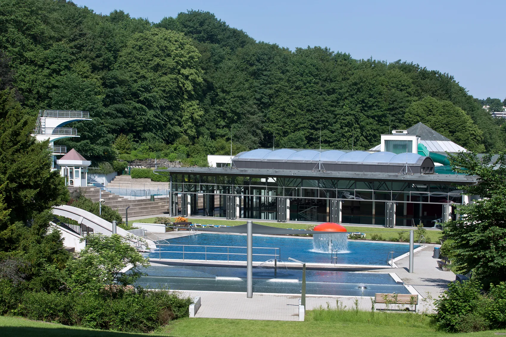 Außenansicht des Freibads Nattenberg mit mehreren Becken, einem Sprungturm und einem modernen Gebäude im Hintergrund, umgeben von Grünflächen.