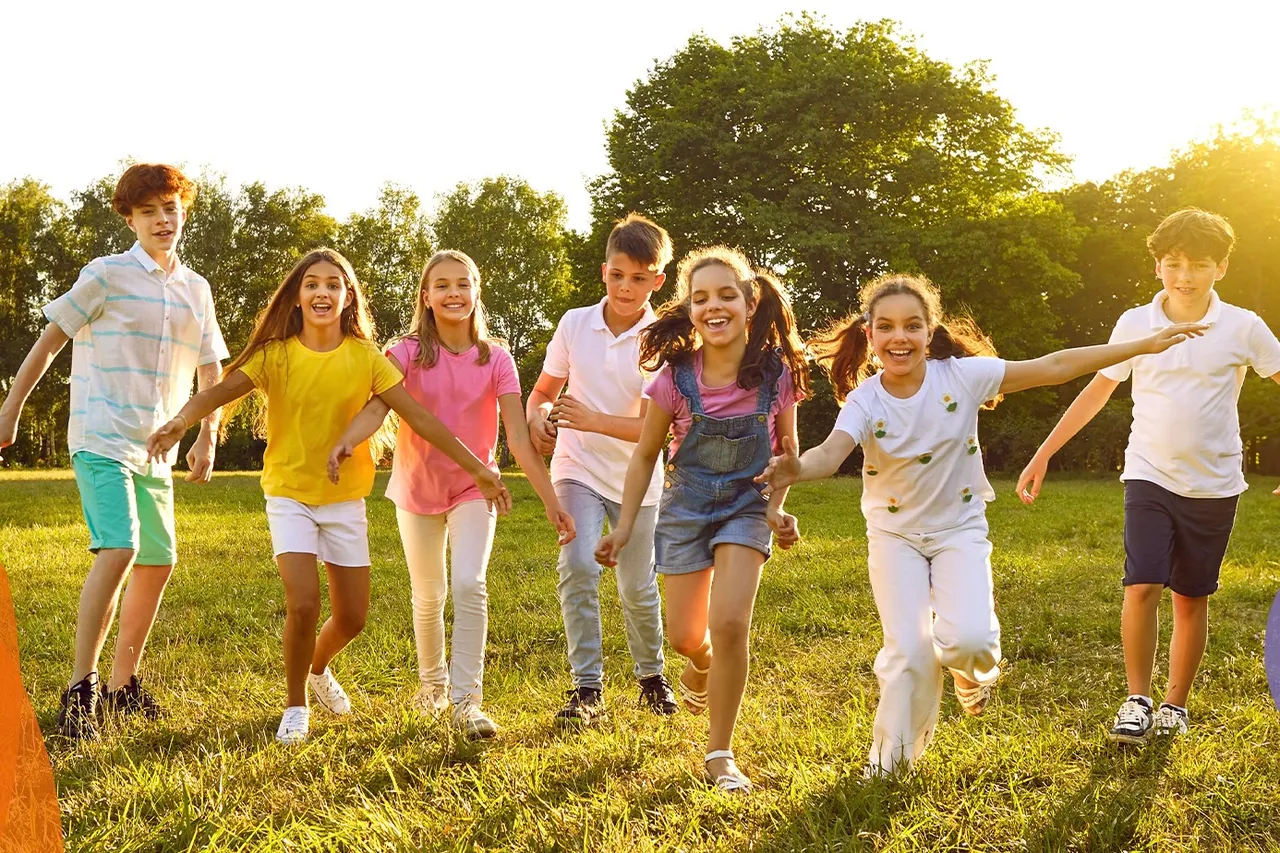 Eine Gruppe von Kindern hält Hände und rennt lachend über eine grüne Wiese in Richtung der Kamera.