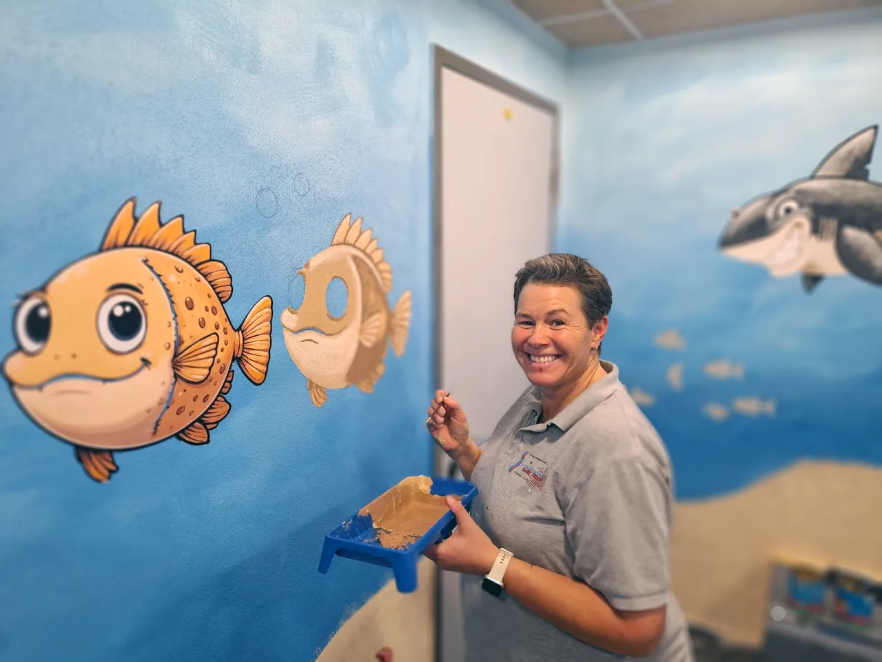 Eine Frau mit graumeliertem Haar lächelt in einem Raum mit einer Wandmalerei von Fischen und einem Hai. Sie hält eine Farbwanne und einen Pinsel.