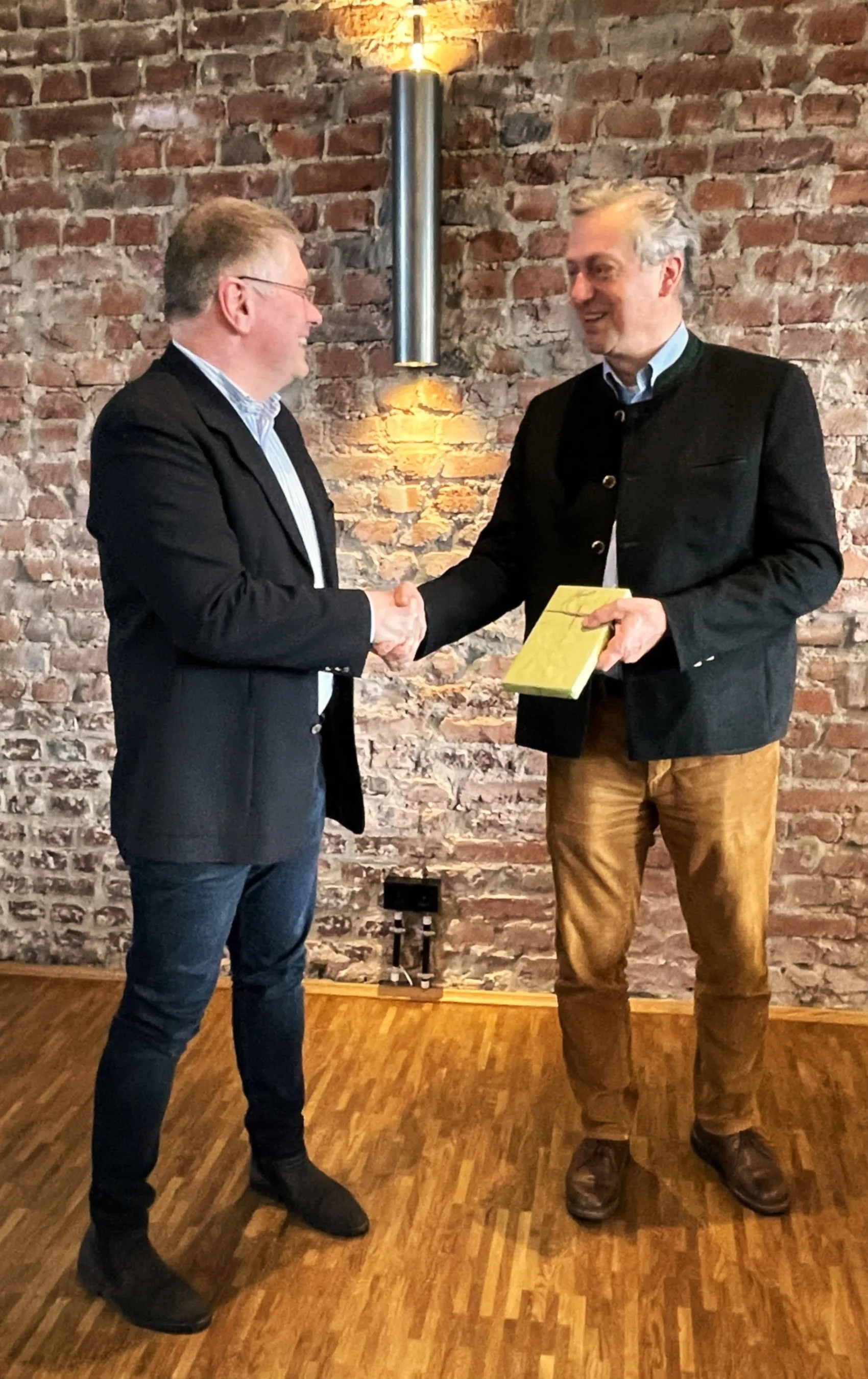 Zwei Männer im Gespräch geben sich die Hand. Der Mann links trägt einen Blazer, der Mann rechts eine Jacke und hält ein Buch.