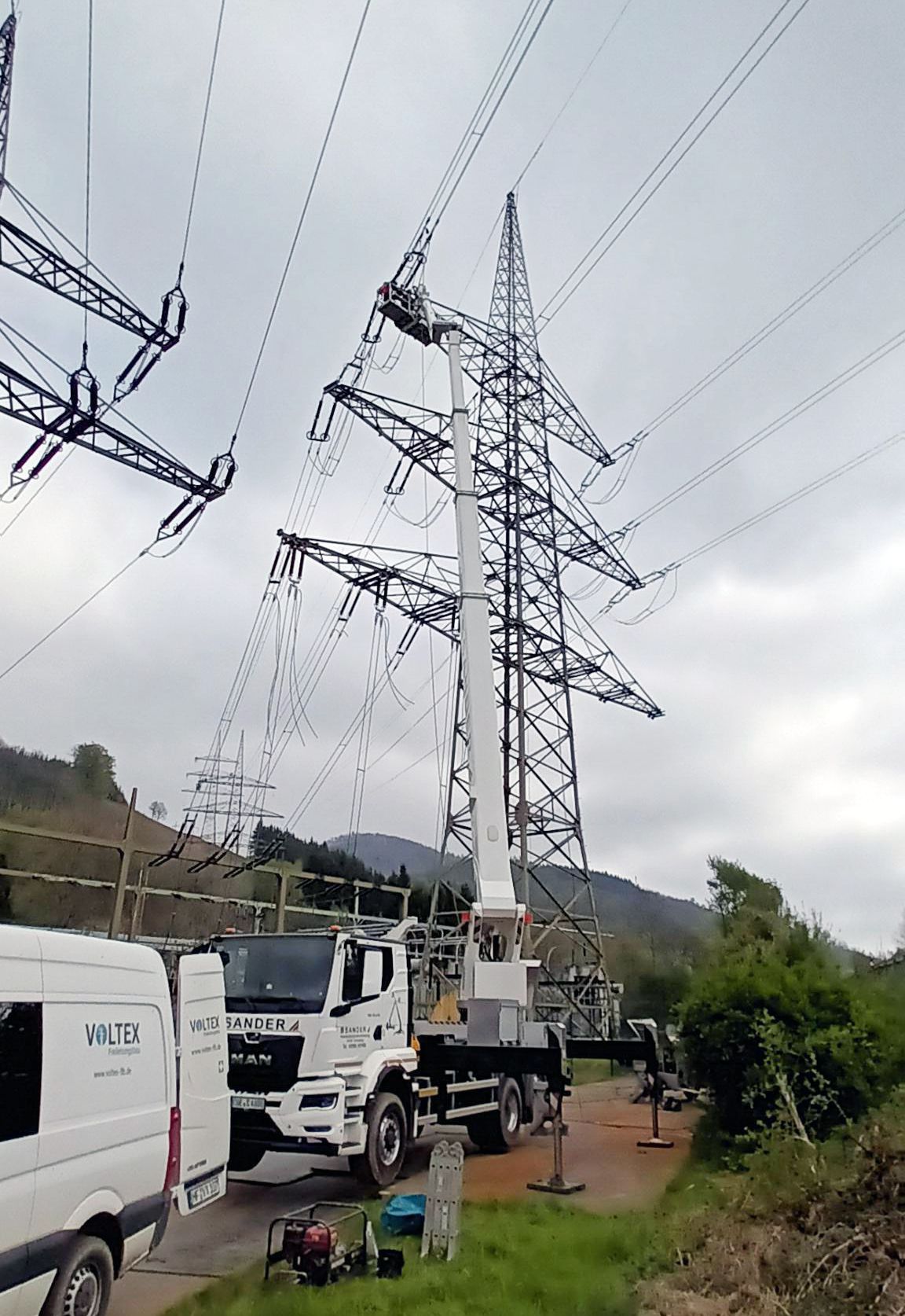 Mehrere Hochspannungsmasten mit neuen Leiterseilen stehen in einer hügeligen Landschaft. Im Vordergrund parken zwei Transporter und ein LKW des Unternehmens Voltex.