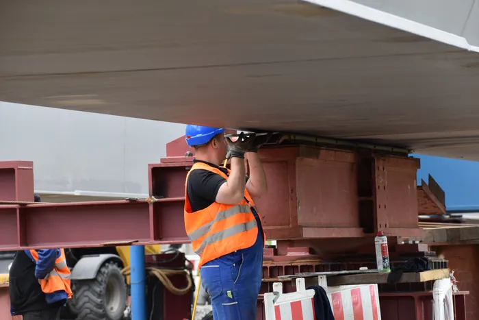 Ein Bauarbeiter in Schutzkleidung und Helm arbeitet unterhalb eines Brückenbauteils, das von einem Transportfahrzeug gehalten wird. Im Hintergrund sind weitere Bauelemente und ein Teil eines Baufahrzeugs zu sehen.