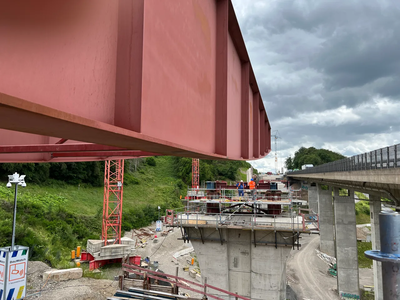 Ein roter Stahlträger wird über eine Brückenbaustelle gehoben. Arbeiter sind auf der Baustelle unterhalb des Trägers zu sehen, während im Hintergrund weitere Brückenstrukturen und ein bewaldeter Hügel zu erkennen sind.