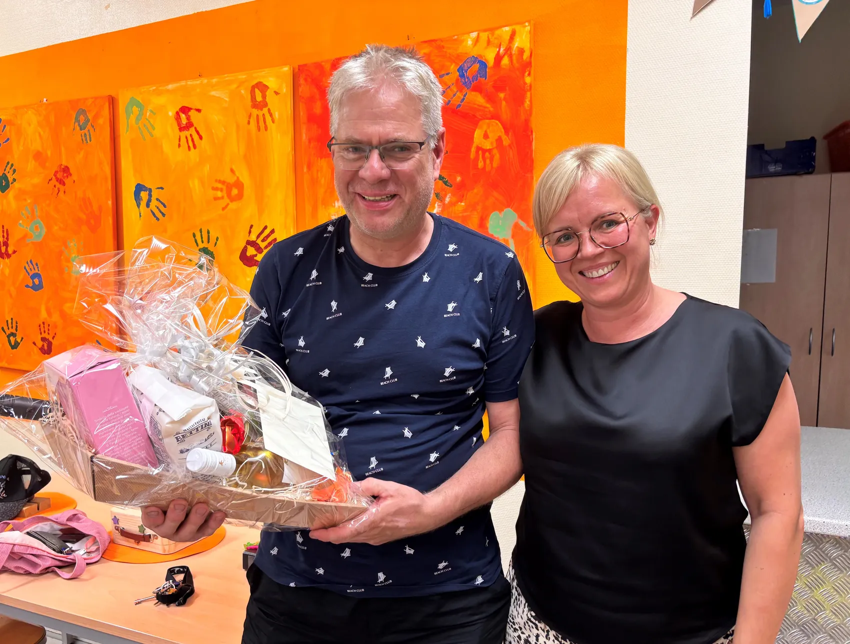 Ein Mann und eine Frau lächeln in die Kamera, zwischen ihnen steht ein Geschenkkorb. Im Hintergrund sind bunte Handabdrücke an einer Wand zu sehen.