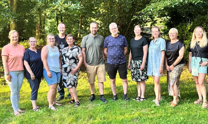 Eine Gruppe von zehn Personen steht auf einer grünen Wiese vor Bäumen. Im Zentrum stehen zwei Männer, von denen der eine ein Hemd mit Muster trägt und der andere ein schwarzes T-Shirt. Die anderen Personen sind Frauen und Männer unterschiedlichen Alters, die lässig gekleidet sind.