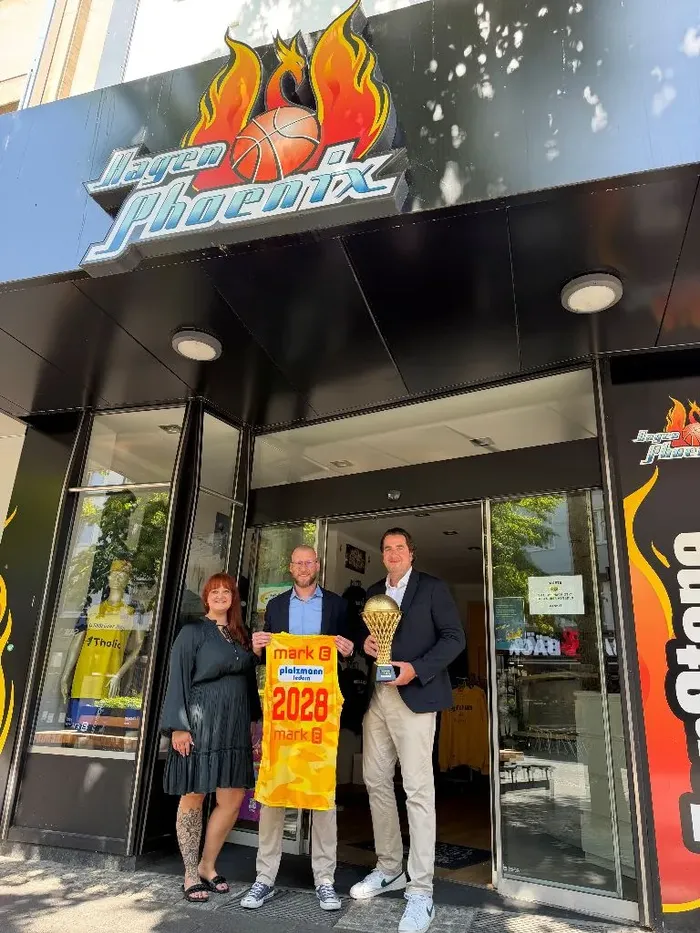 Kira Schurna, Martin Schmidt und Alexander ten Hompel halten ein gelbes Banner mit „2028“ und einen Pokal vor dem Eingang von Phoenix Hagen.