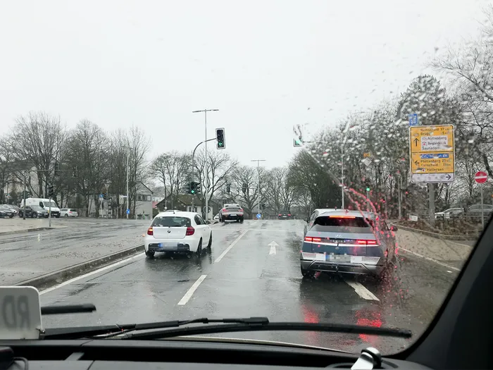 Blick durch eine regennasse Windschutzscheibe auf eine Straße mit fahrenden Autos und Ampeln. Im Hintergrund sind Schilder mit Ortsangaben zu sehen.