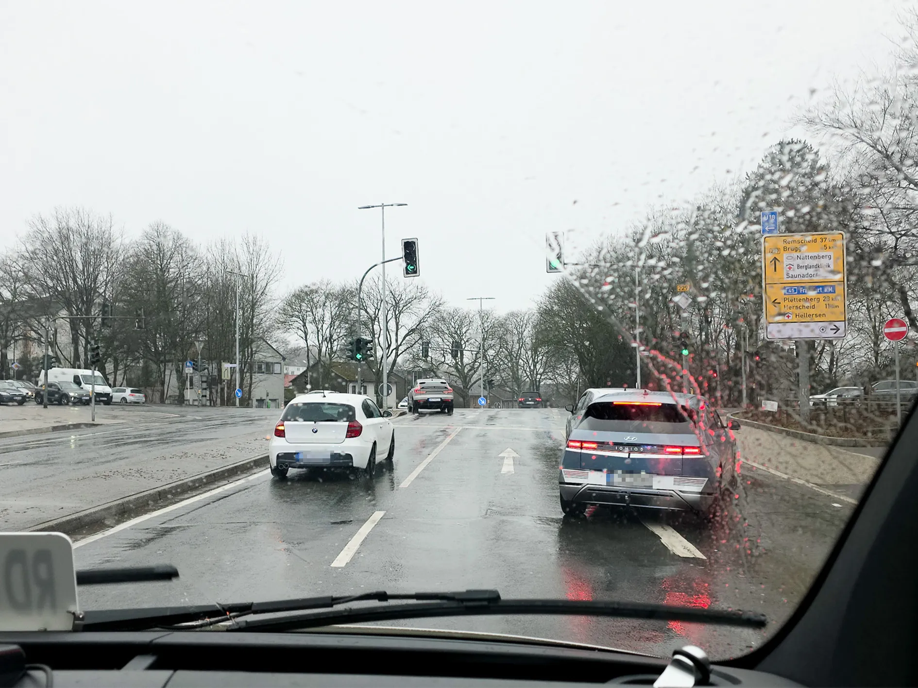 Blick durch eine regennasse Windschutzscheibe auf eine Straße mit fahrenden Autos und Ampeln. Im Hintergrund sind Schilder mit Ortsangaben zu sehen.