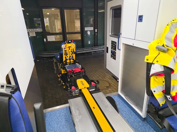 Ein gelber Tragenlift steht in einem Zugwagen bereit, um eine Trage nach draußen zu fahren. Ein Rettungssanitäter in gelber Schutzkleidung steht daneben.