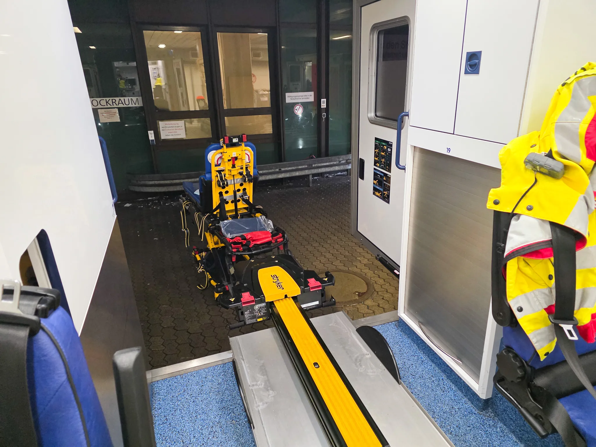 Ein gelber Tragenlift steht in einem Zugwagen bereit, um eine Trage nach draußen zu fahren. Ein Rettungssanitäter in gelber Schutzkleidung steht daneben.