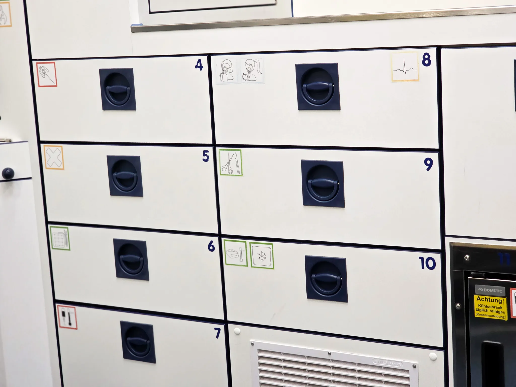 Weißer Schrank mit mehreren Fächern, jeweils mit einem runden Griff und einem Piktogramm versehen, vermutlich zur Kennzeichnung des Inhalts.