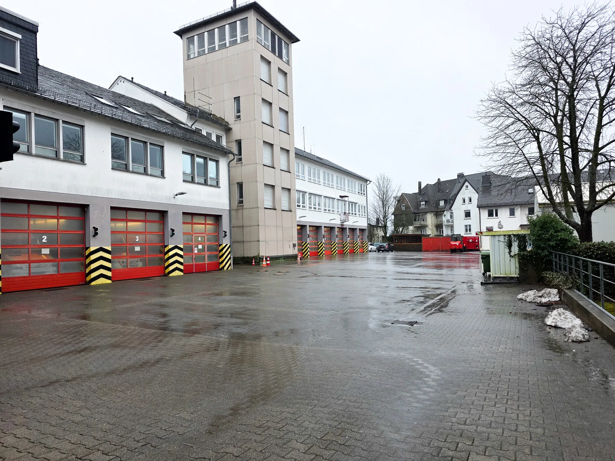 Das zweigeschossige Feuerwehrhaus mit roten Rolltoren und einem Wachturm steht vor einer Wohnbebauung. Vor dem Gebäude befindet sich ein nasser Hof.