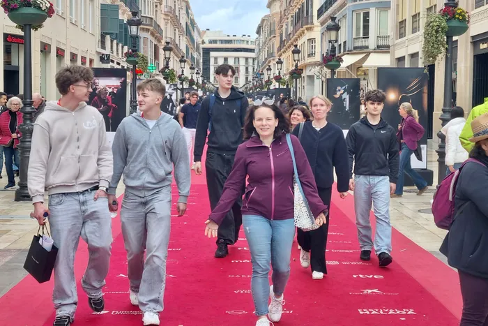 Eine Gruppe junger Menschen geht auf einem roten Teppich durch eine belebte Straße in Málaga, Spanien. Im Hintergrund sind Gebäude und Geschäfte zu sehen.