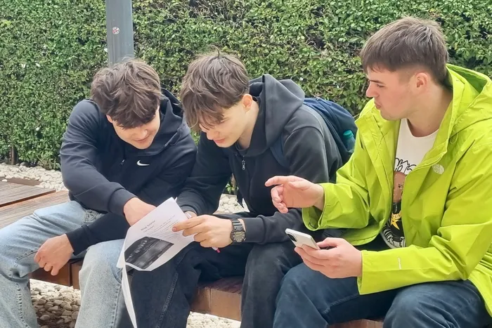 Drei junge Männer sitzen im Freien zusammen und betrachten ein Dokument sowie ein Smartphone. Sie absolvieren ein berufliches Praktikum in Málaga.
