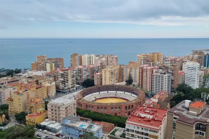 Luftaufnahme von Málaga, Spanien, zeigt eine Stierkampfarena inmitten dichter Wohnbebauung und im Hintergrund das Mittelmeer.
