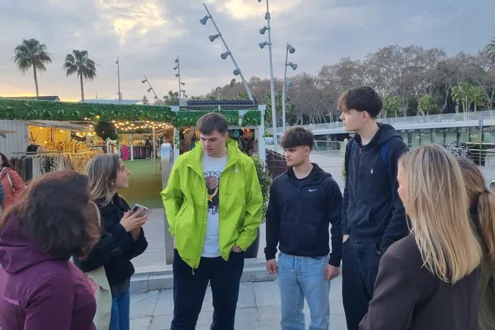 Eine Gruppe junger Menschen steht auf einem Platz vor Marktständen in Málaga, Spanien. Ein Schüler filmt die anderen mit einem Smartphone.