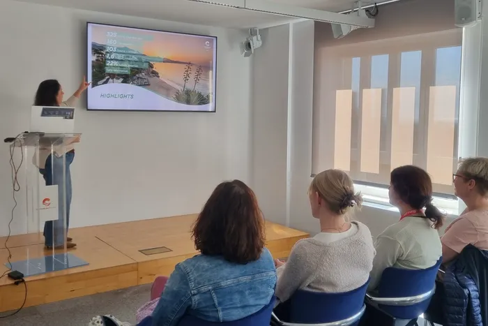 Eine Frau präsentiert vor einem Publikum eine Präsentation mit Statistiken und einem Bild einer Küstenlandschaft. Im Hintergrund sind orangefarbene Säulen und eine helle Decke zu sehen.